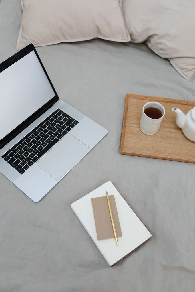 From above of book with jotter and pen arranged on comfortable bed with opened laptop and tray with coffee cup and pot during freelance work