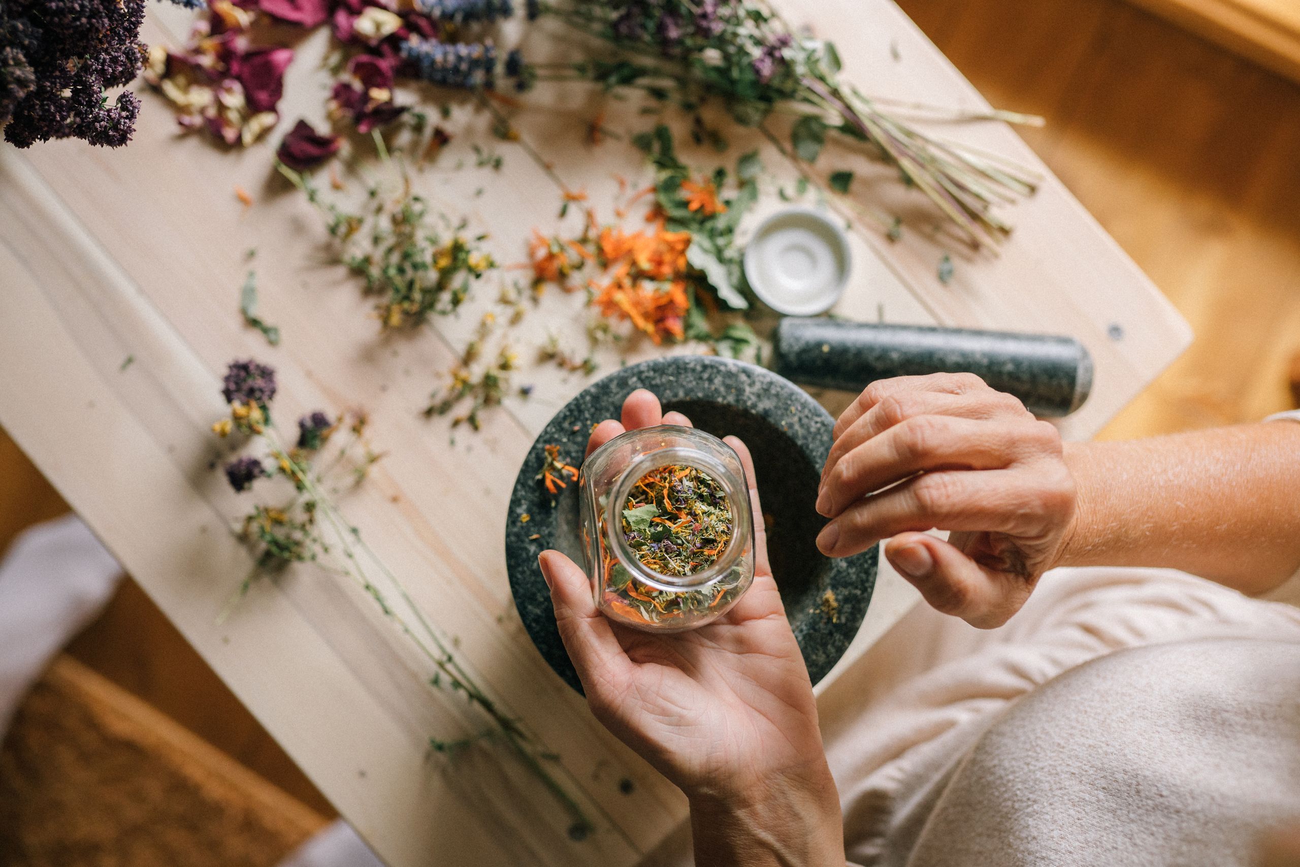 Top view of hands preparing herbs and dried flowers using a mortar and pestle on a wooden table.